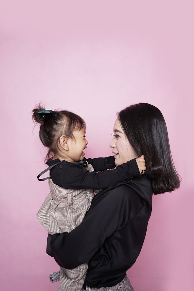A happy mother holding her smiling daughter against a pink backdrop, showcasing love and togetherness.