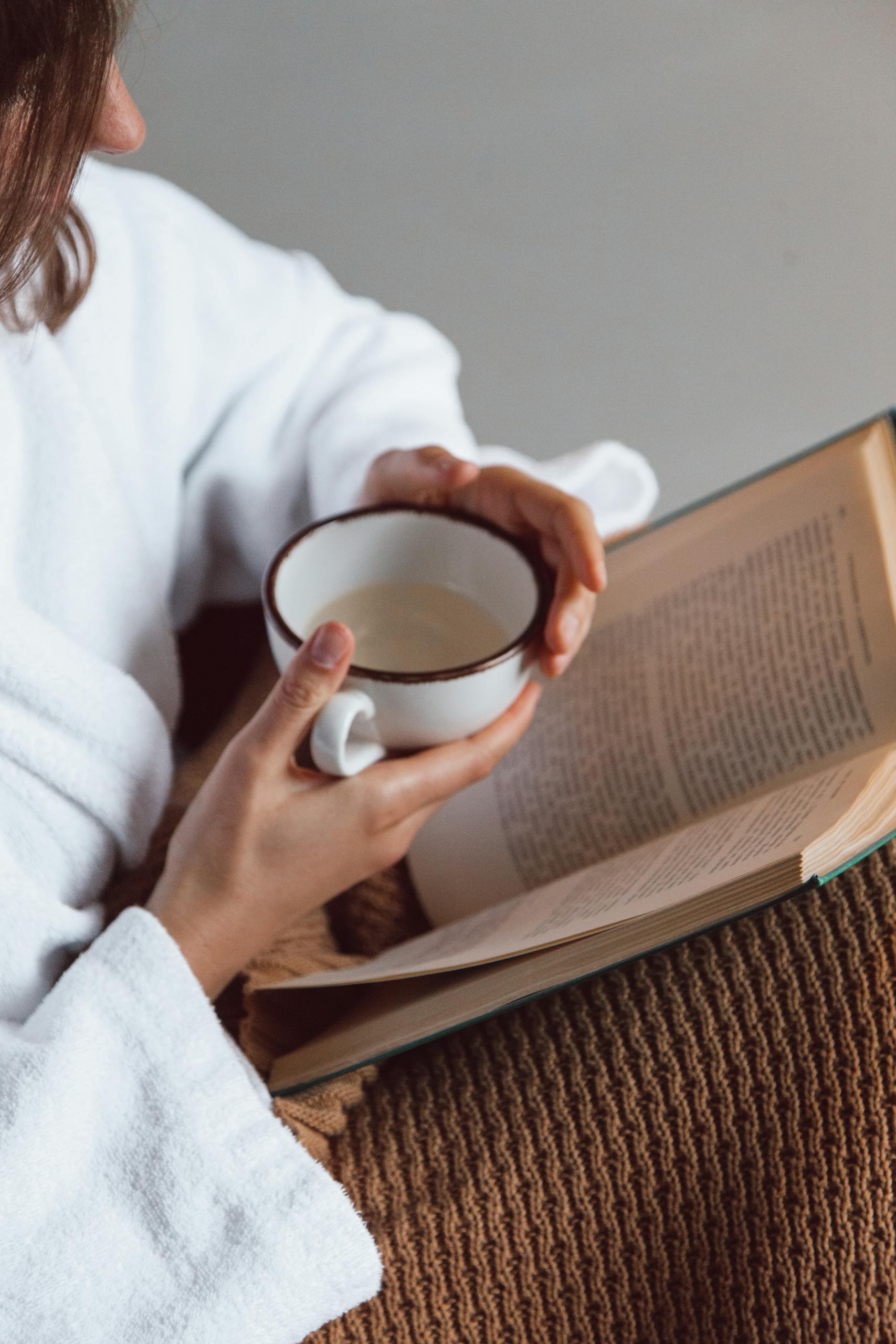 A person enjoying a cozy moment with a book and a cup of tea in hand, wearing a robe.