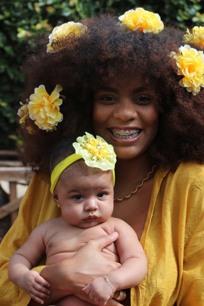 A smiling mother with floral hair decor holds her baby outdoors, creating a joyful family scene.