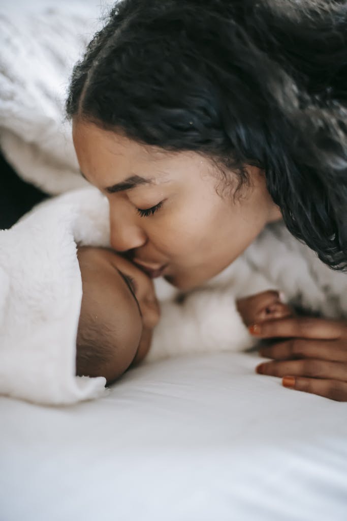 Calm ethnic mother kissing little baby lying on bed and sleeping at home in daylight