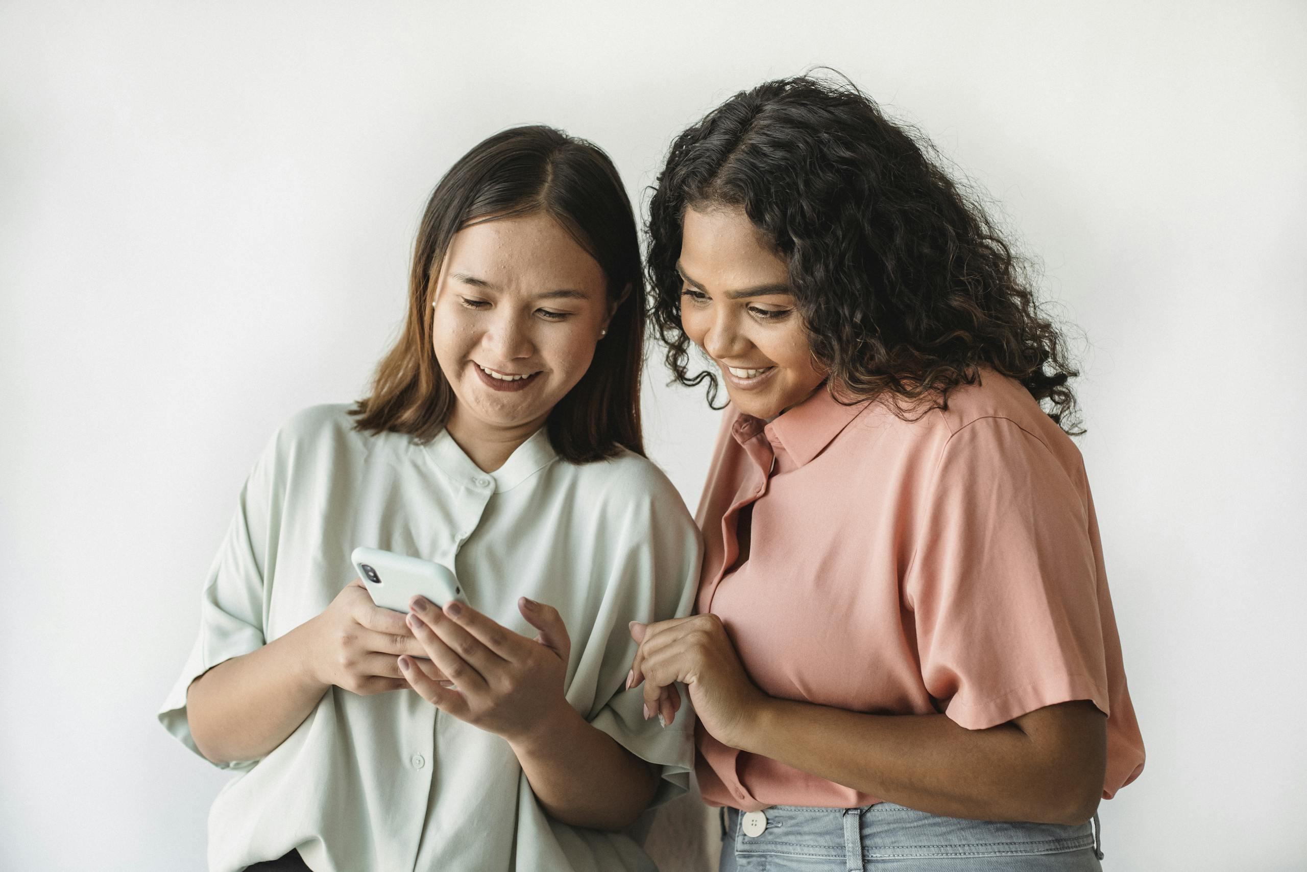 Two women smiling and looking at a smartphone, sharing a joyful moment indoors.